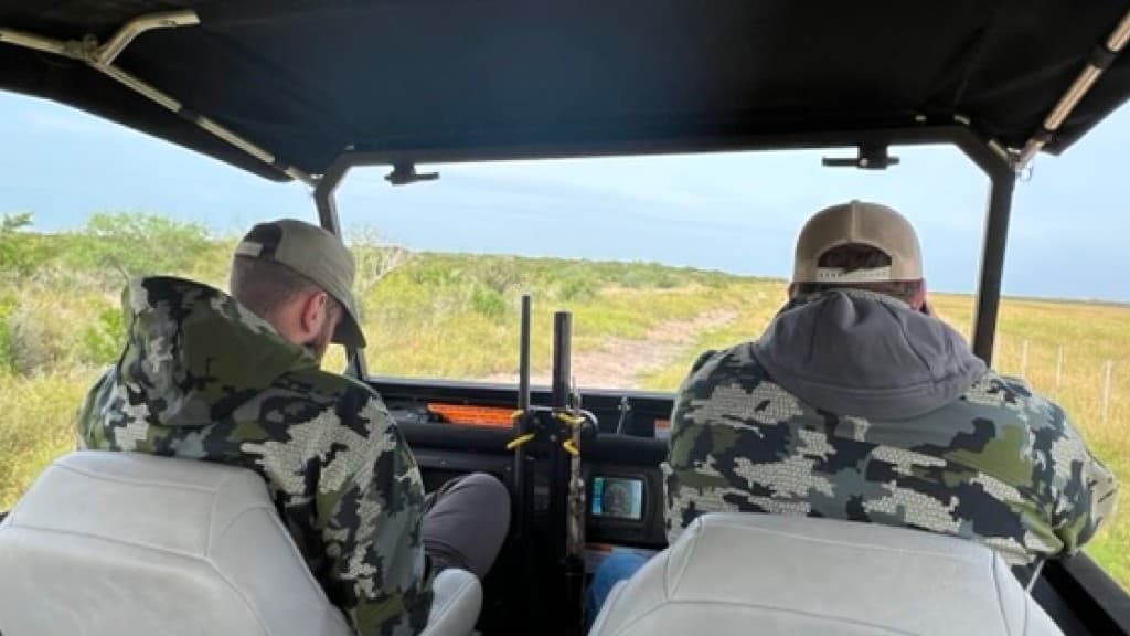 Two hunters in camo riding in a side-by-side looking out over open ranch land